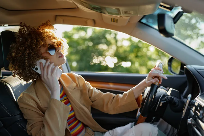 Cheerful woman driving a car, talking on the phone, curly hair and sunglasses, sunlit interior, retro film color, casual fashion, lifestyle moment, candid outdoor scene