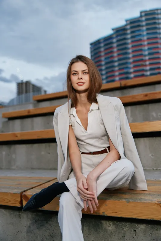 confident woman in white business outfit sitting on urban wooden stairs under cloudy sky with modern building background