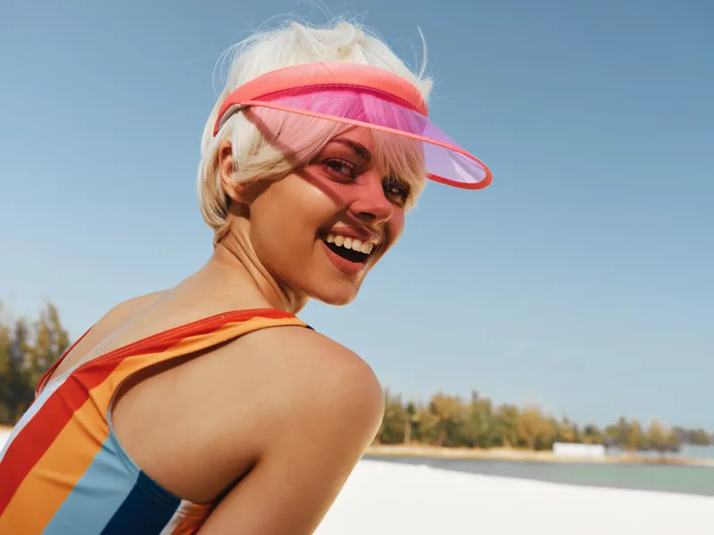 Bright sunlit portrait of a smiling woman at the beach wearing a colorful striped swimsuit and pink visor, capturing playful summer energy and carefree outdoor vibes.