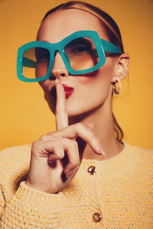 Stylish woman in large turquoise sunglasses and yellow sweater makes shushing gesture with finger on lips. Close-up portrait on yellow background. Concept of silence, secrecy, and fashion.