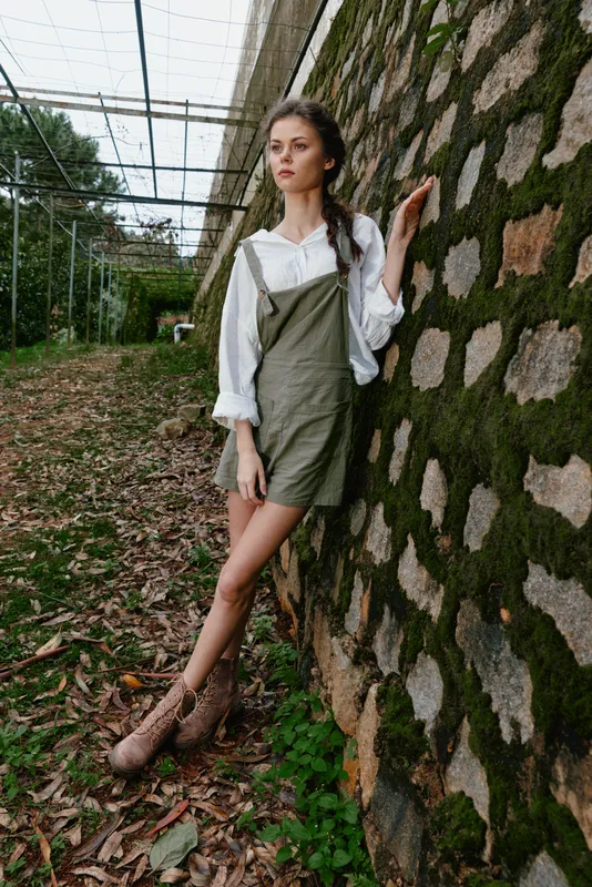 Young woman in casual green dress and white shirt posing against a textured stone wall, reflecting a relaxed, natural outdoor lifestyle in a summer setting