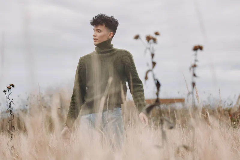 Candid portrait of a young man walking through tall grass in a textured sweater, shot with soft natural light and muted tones to convey genuine emotion, authenticity and credibility.