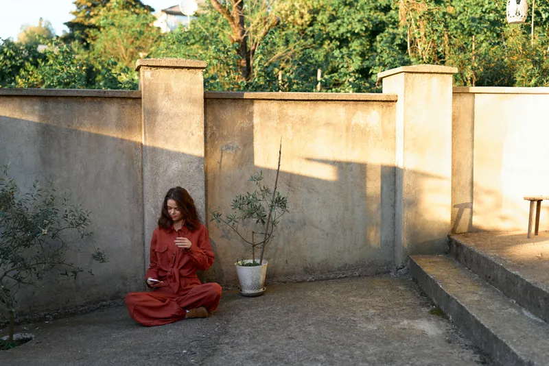 A woman in a red outfit sits cross legged on a sunlit courtyard, leaning against a weathered concrete wall, quietly contemplating solitude and inner calm