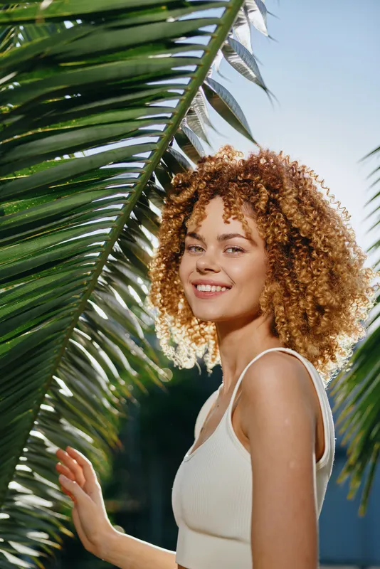 smiling woman with curly hair enjoying sunlight outdoors near tropical palm leaves, wearing white top and expressing happiness on a bright sunny day