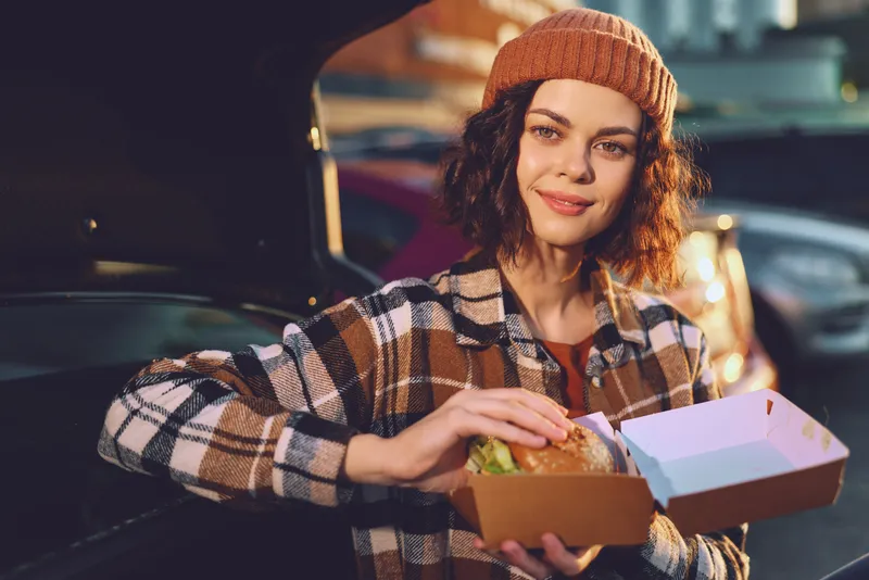 Woman holding burger in takeout box by car trunk, beanie and plaid jacket, smile and candid lifestyle moment with authenticity and golden hour glow, urban mindful living vibe.
