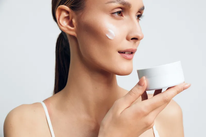Young woman applying skincare cream to face, holding a small cosmetic cup, focused profile, smooth skin, beauty spa hygiene mood, bright studio light, white background