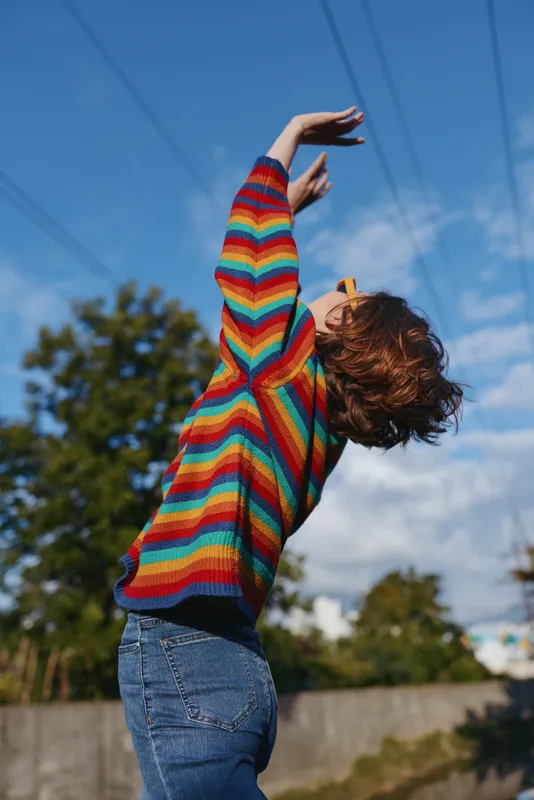 Woman in colorful striped sweater outdoors wearing sunglasses, joy and freedom as she stretches her arms up toward blue sky. Casual fashion, motion and playful energy.