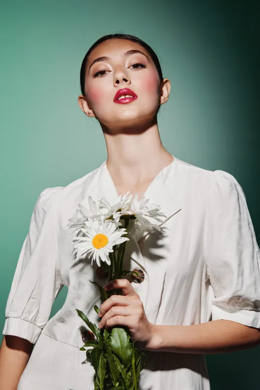 Young woman in a white dress holds fresh white daisies against a green background. Her makeup is vibrant and artistic with glossy lipstick and defined eyebrows creating a fashion beauty concept.