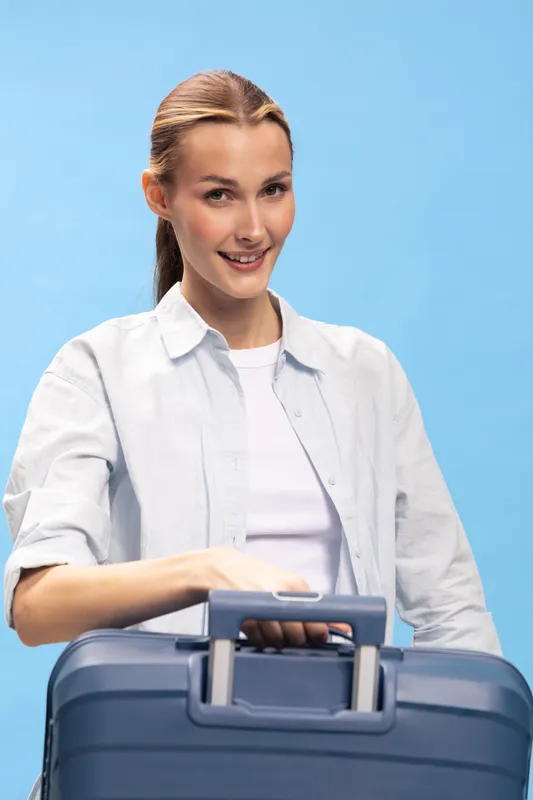 Smiling woman with tied hair holds blue suitcase on blue background. Casual clothing and travel concept with happiness and positive mood for trip or vacation.