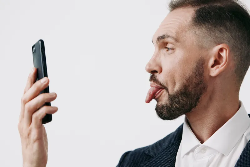 A professional man in a suit playfully sticks out his tongue toward a smartphone, conveying curiosity, humor, and bold attitude against a clean solid background.