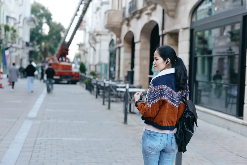 Street scene featuring a woman in a knit sweater and backpack on a city sidewalk, distant construction crane in the background, a candid urban fashion moment.