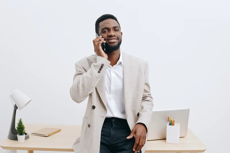 Professional young African American man in smart casual attire holding a phone and smiling while standing in a modern office environment with a desktop computer, plant, and stationery, white backgroun