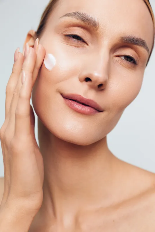 Smiling woman applying white face cream, close up on cheek, soft studio lighting, clean light background, skincare hydration wellness self care, beauty routine for healthy skin