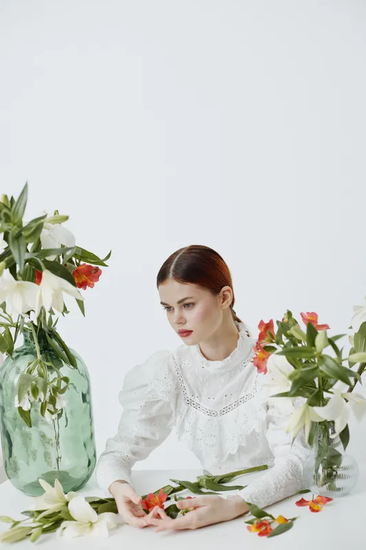 A pensive model in a vintage white dress surrounded by elegant flowers, expressing serene emotions in a minimalist studio setting