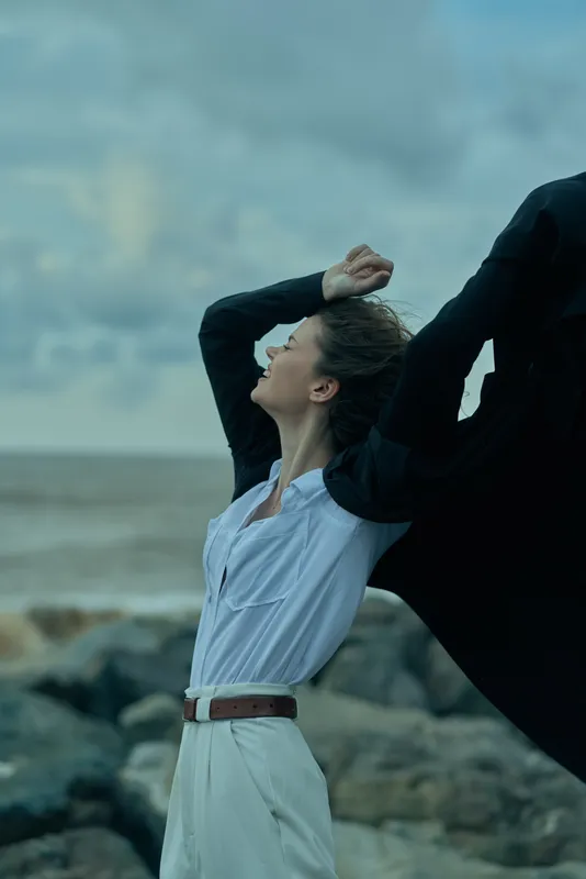 Woman on rocky shore lifts a flowing cape against the wind, white belt dress billowing, dramatic coastal scene highlighting movement, fashion, drama and natural light by the sea