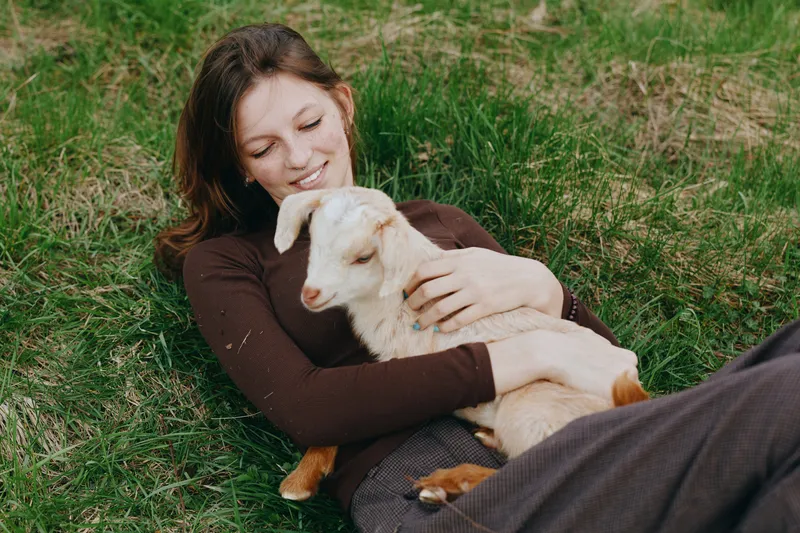 A woman lies on the grass, smiling as she cuddles a playful goat kid in her arms, surrounded by sunlight and greenery. A peaceful lifestyle moment emphasizing bond between human and animal outdoors.