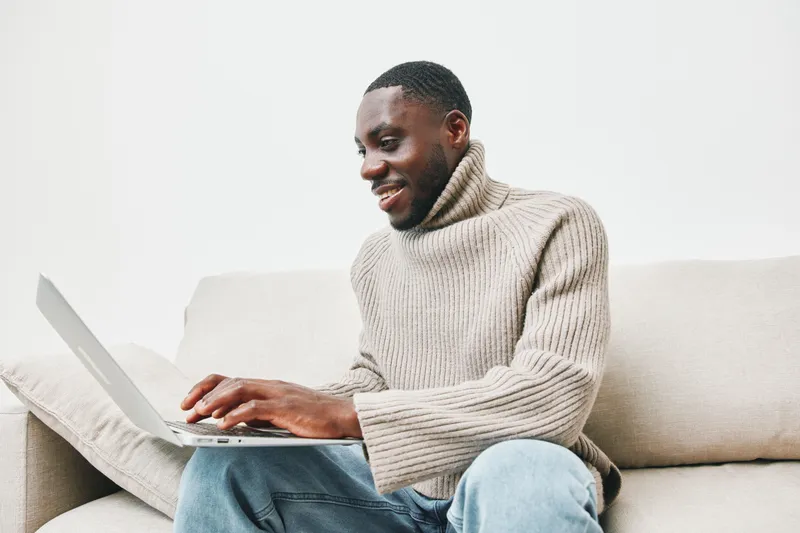 Young Black man smiling while working on a laptop at home in a cozy sweater, capturing a joyful and relaxed atmosphere