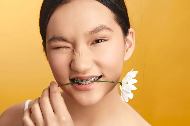 asian woman with a bright smile holds a white flower between her lips, posing against a warm orange backdrop to convey playful charm and gentle creativity in a studio portrait.