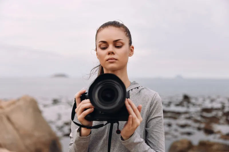 Young woman holding a camera by the coast in a sporty outfit, showcasing confidence and a passion for photography in a serene coastal setting