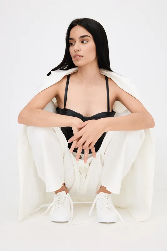 young woman in white pants and black top squatting thoughtfully against white background, modern style, casual sneakers, natural look