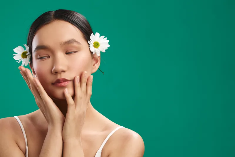 Asian woman with floral accessories rests her face softly in her hands against a green backdrop, conveying calm beauty and a gentle, meditative mood in a clean studio setting.