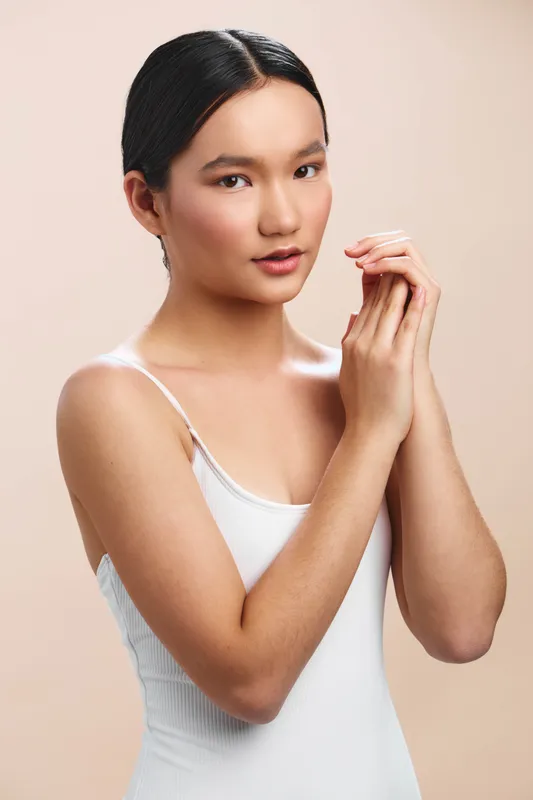 asian woman stands in a studio wearing a white tank top, soft lighting highlights her smooth skin and serene expression, creating a clean, elegant portrait with a confident, modern vibe.