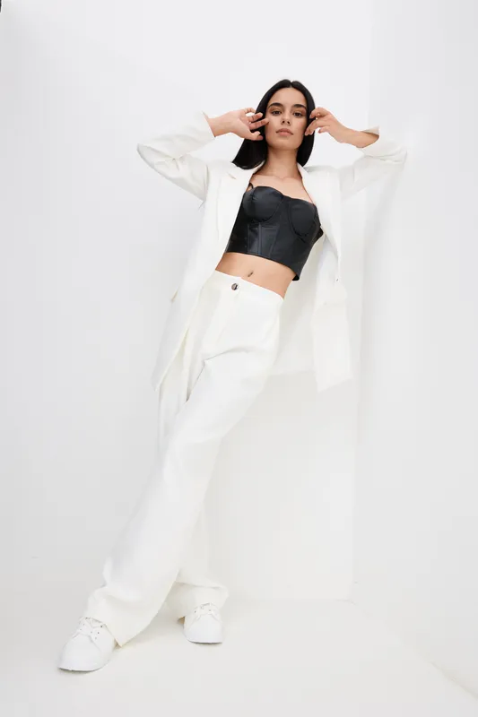 Stylish woman in white pantsuit and black bustier poses confidently against minimal white background, showcasing modern fashion and monochrome elegance.