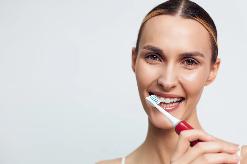 Young woman with natural skin smiles while brushing her teeth with a red toothbrush isolated on white background, showing healthy dental care and personal hygiene routine.