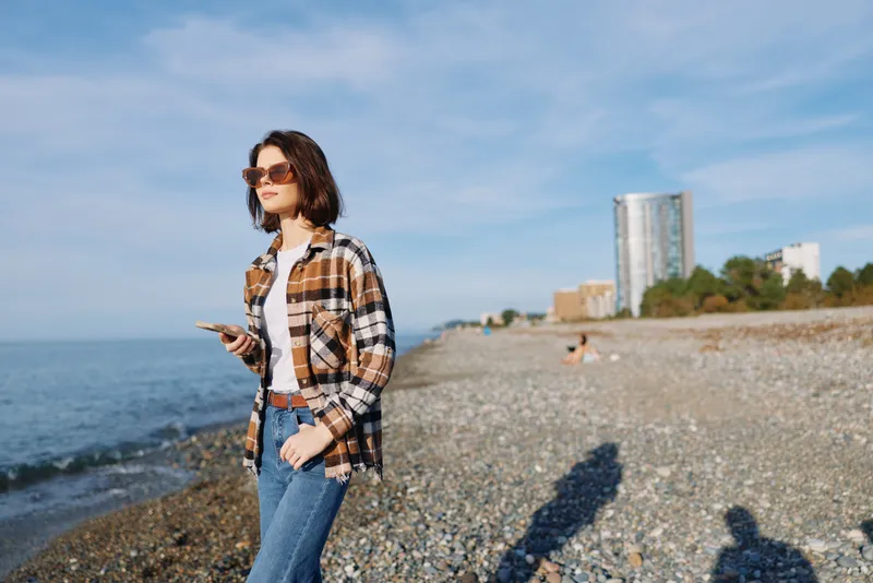Woman on a sunny beach walks along the shore, wearing a plaid shirt and jeans, glancing at her phone with sunglasses, distant city buildings and a bright blue sky overhead.