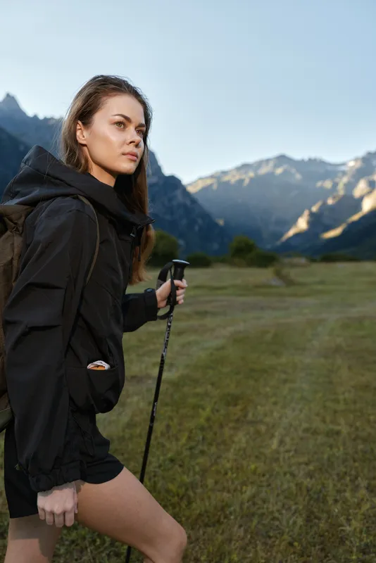 A focused hiker strides across a grassy meadow with a trekking pole in hand. She wears a black jacket and shorts, while distant mountains and a clear sky frame a rugged, adventurous outdoor scene.