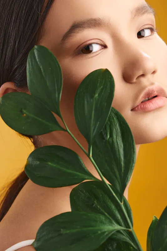 asian woman with silky skin gazes softly while large leaves frame her face in a studio portrait, creating a fresh natural mood and elegant contemporary aesthetic.