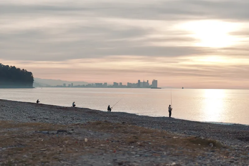 Beach at sunset with silhouetted figures along the shore, calm water, distant city skyline on the horizon, scattered pebbles, gentle waves, and a peaceful vibe perfect for travel, nature, and