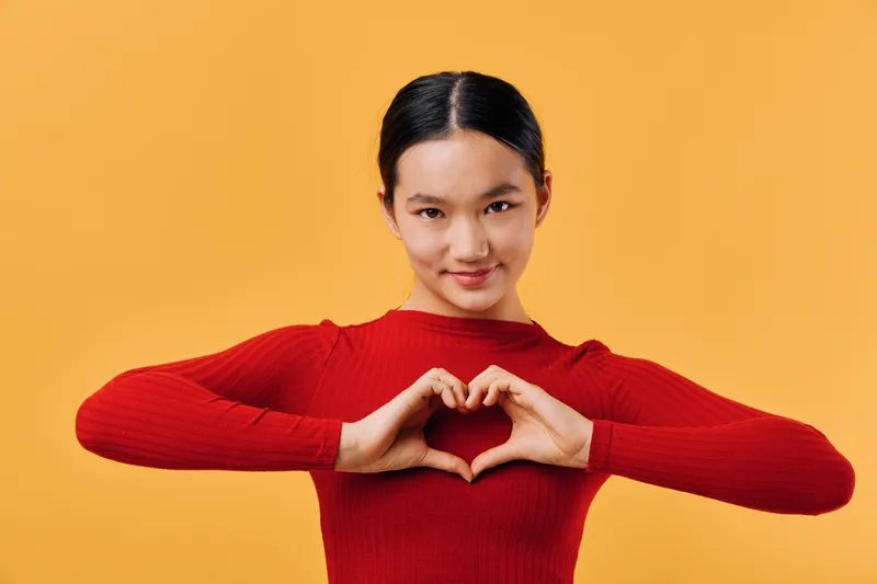 Young woman in red sweater making heart shape with hands and smiling on orange background. Gesture for love, positivity and connection with cheerful expression and casual look.