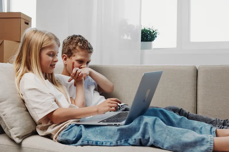 Two children sitting on a sofa using a laptop together in a bright room. Casual clothing and relaxed atmosphere with natural light.