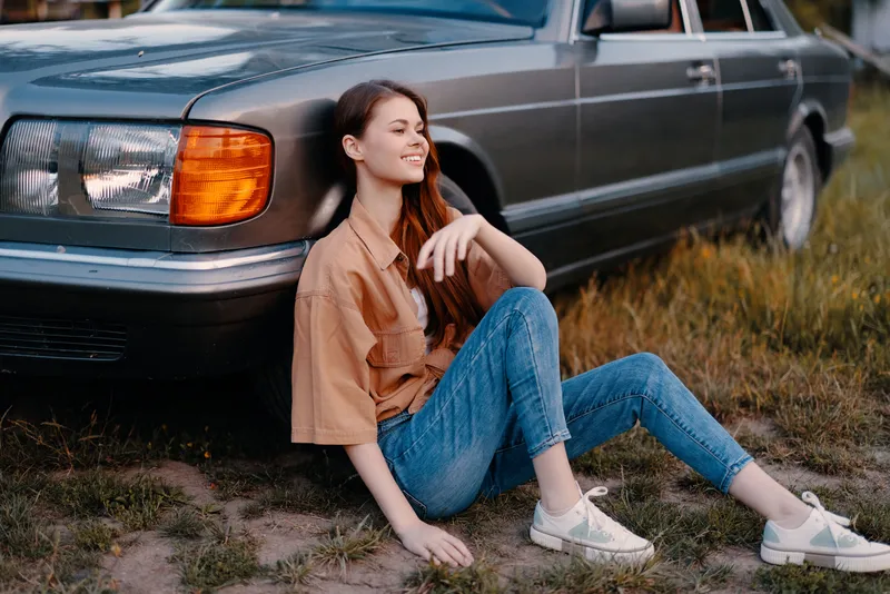 Young woman sitting beside vintage car, smiling in casual outfit, summer vibe with green grass background, perfect for lifestyle and travel themes.