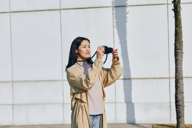Photography of a casual photographer outdoors, wearing a beige coat and denim, raising a camera to capture a candid shot under bright daylight.