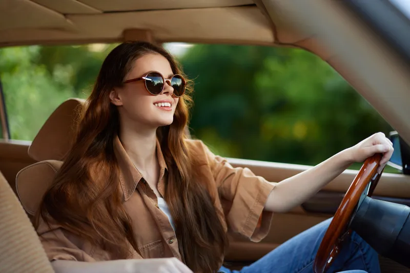 Young woman driving a vintage car in stylish sunglasses, smiling joyfully in a sunny outdoor setting, evoking a sense of freedom and adventure.
