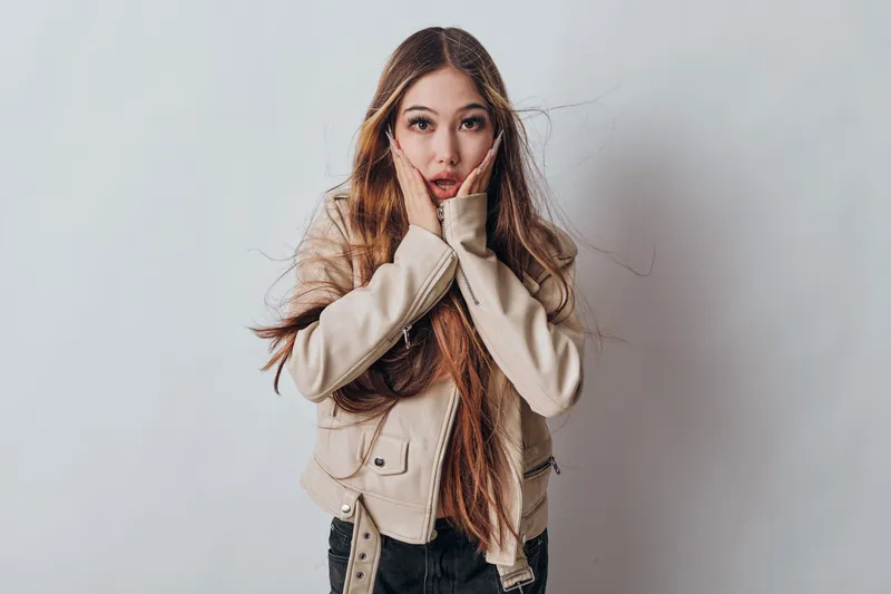 Young woman with long hair in beige jacket looks surprised or shocked with hands near face in studio on light background for emotional portrait concept.