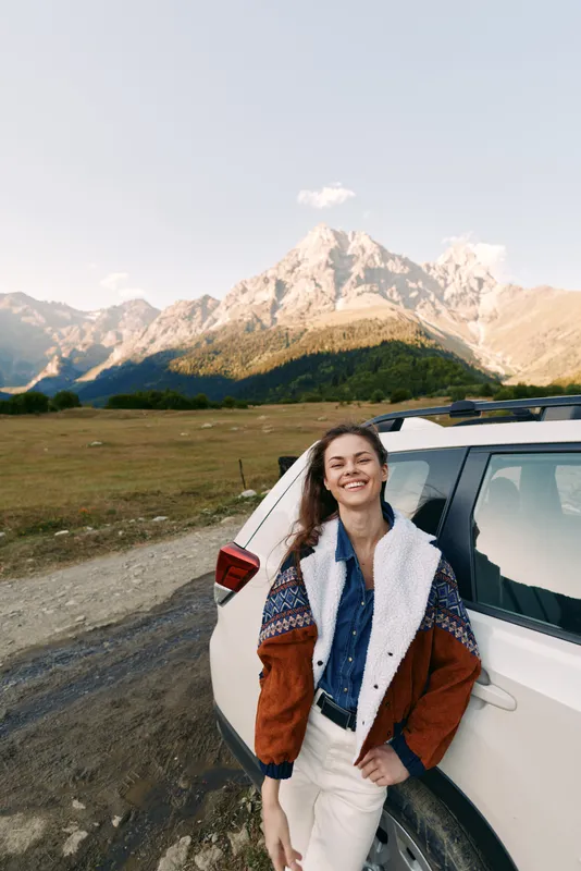 Woman leaning on car by mountains landscape, smile at camera during travel. Nature scene with road and SUV, relaxed portrait, outdoor adventure and scenic daylight vibes.
