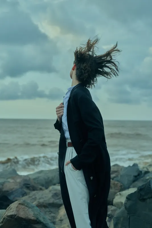 Windy coastal scene with a woman in a blazer and white pants facing the sea, hair and coat blown by wind, dramatic sky over rocky shore