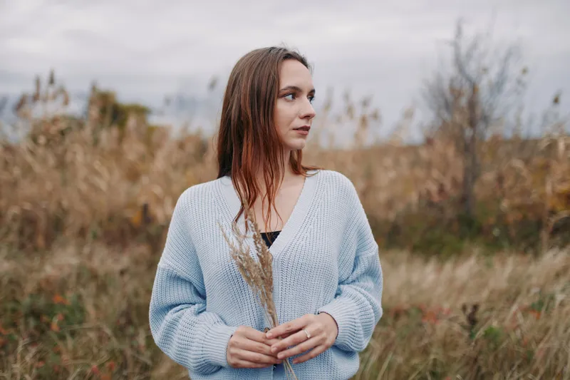A genuine, candid portrait of a young woman in a meadow wearing a cozy sweater, captured in natural light to convey honest, true-to-life emotion and quiet contemplative focus.