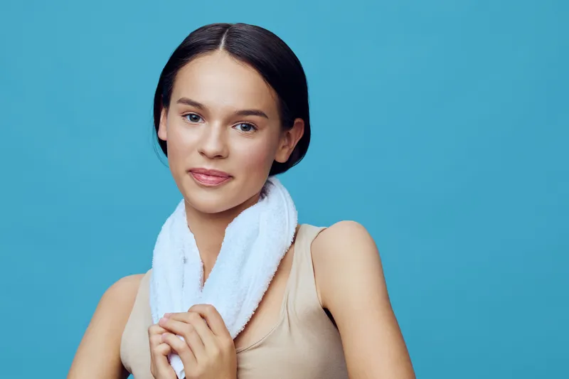 A young woman with a towel around her neck, showcasing a healthy lifestyle and a confident smile against a blue background, reflecting wellness and self care