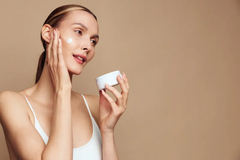 Young woman applying face cream with fingers, skincare routine, natural beauty, soft lighting, neutral background, healthy glowing skin, casual white top