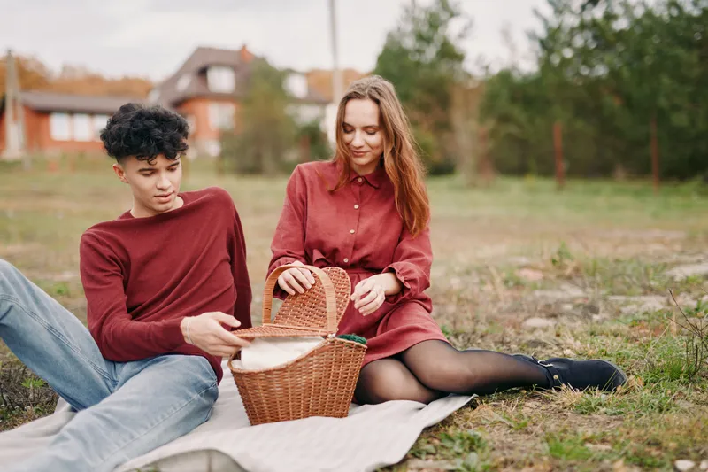 Authentic moment of two friends outdoors sharing a simple picnic, candid expressions, warm tones, casual outfits, genuine connection, natural setting, soft daylight.
