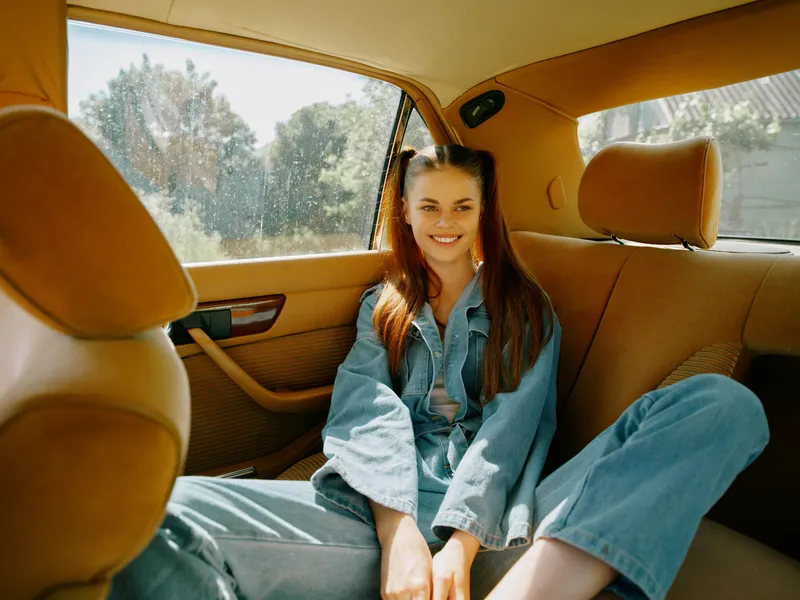 Young woman in casual denim outfit sitting in a warm-colored car, smiling, representing joy and freedom, perfect for lifestyle and travel themes.
