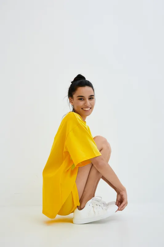 smiling woman in bright yellow oversized t-shirt and white sneakers sitting crouched against white wall with casual hairstyle and relaxed expression