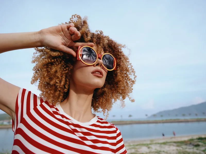 Portrait of a young woman with curly hair wearing a red striped shirt and round sunglasses, posing confidently on a sunny beach with calm water and distant hills.
