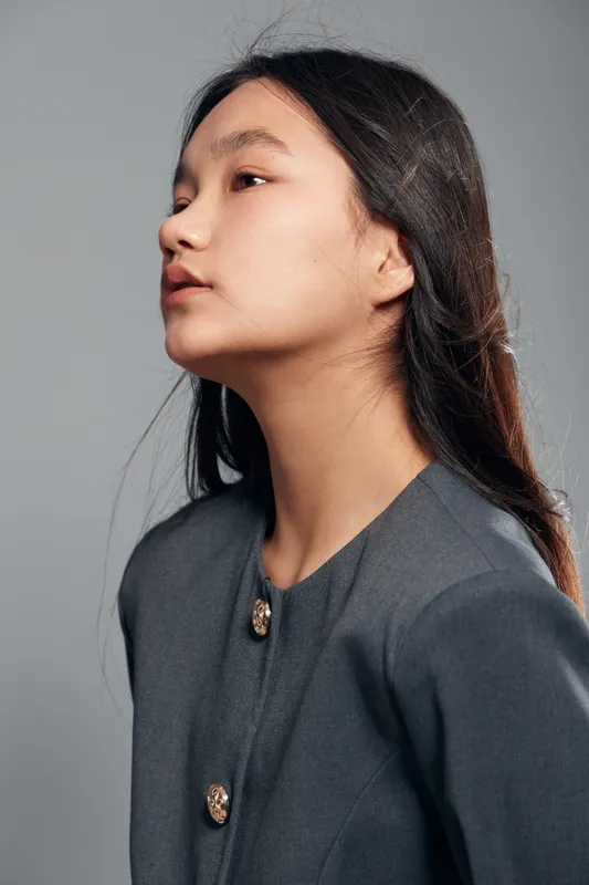 Portrait of young woman with long black hair and grey jacket looking sideways against light grey background. Minimal makeup and natural expression with soft studio lighting.
