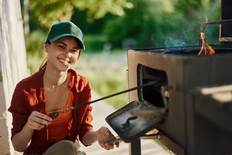 Happy young woman grilling outdoors in summer, wearing casual clothing and a cap, enjoying a sunny day with vibrant greenery in the background