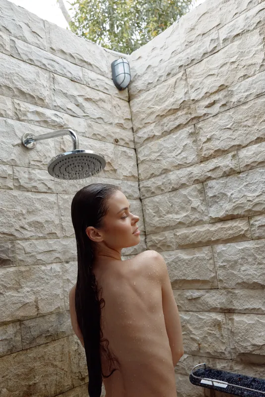 Young woman enjoying outdoor shower with wet hair against natural stone wall in daylight, relaxed and peaceful moment in summer atmosphere.